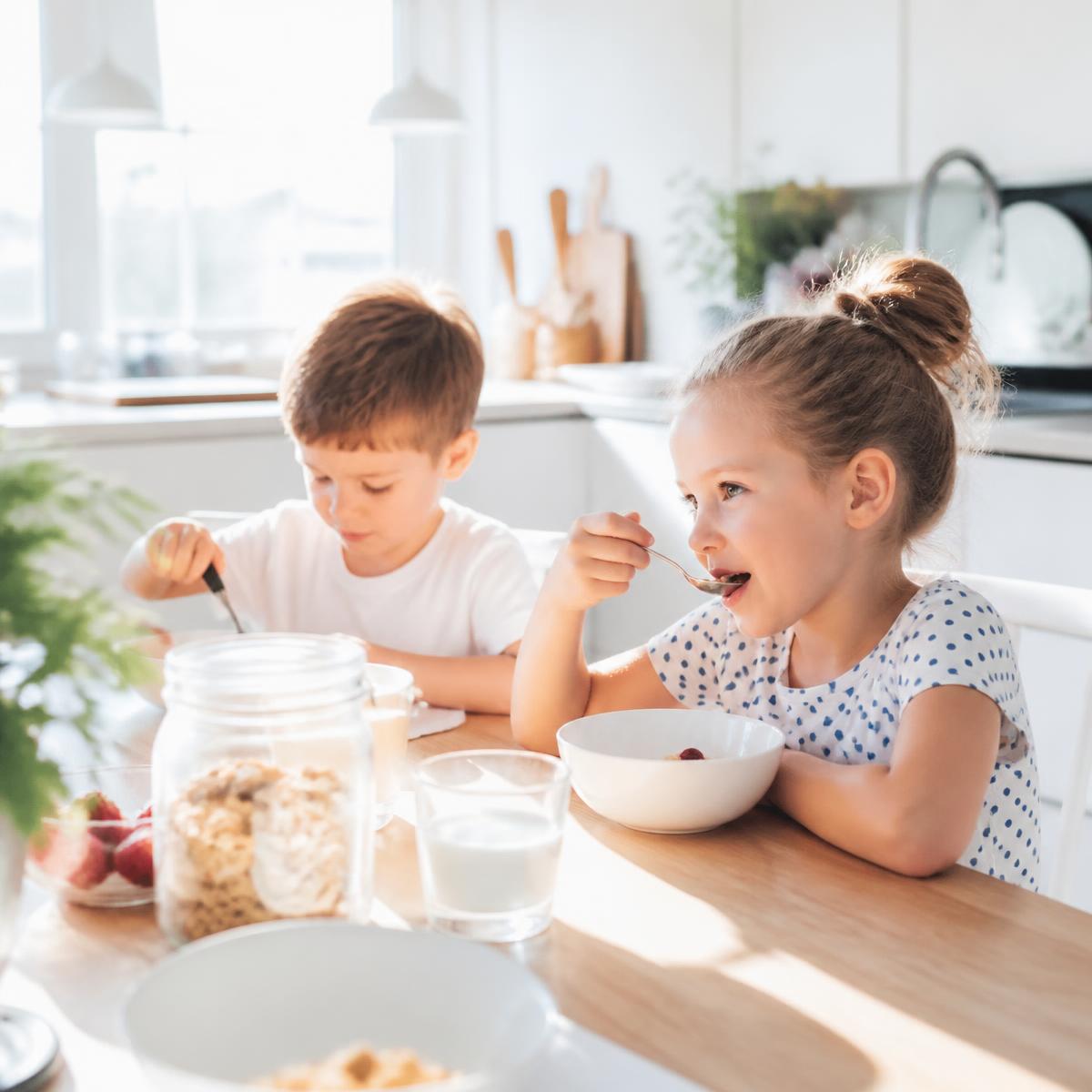 Two young children eating cereal at a kitchen counter in morning light