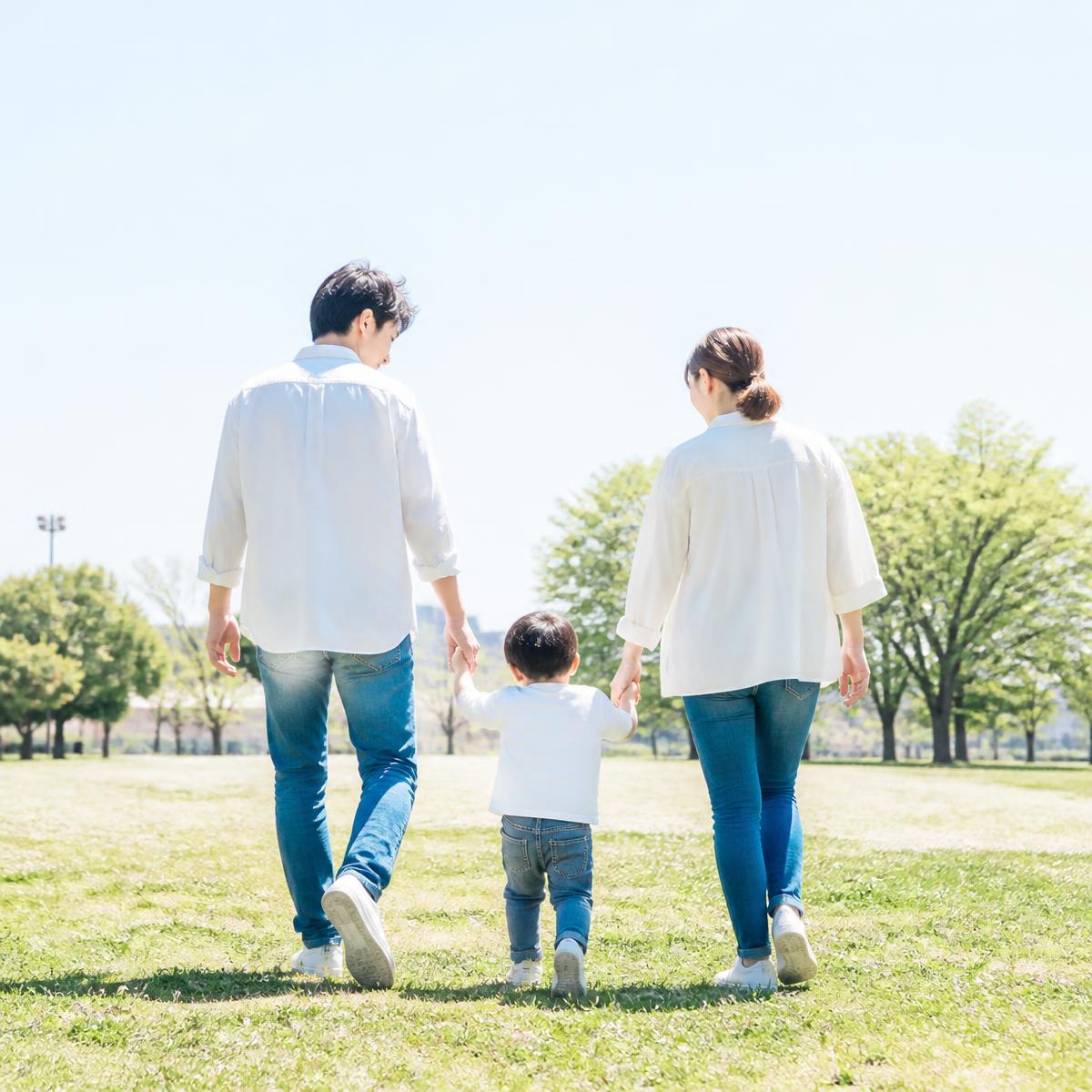 Two parents seen from behind walking across sunny parkland holding their toddler’s hands